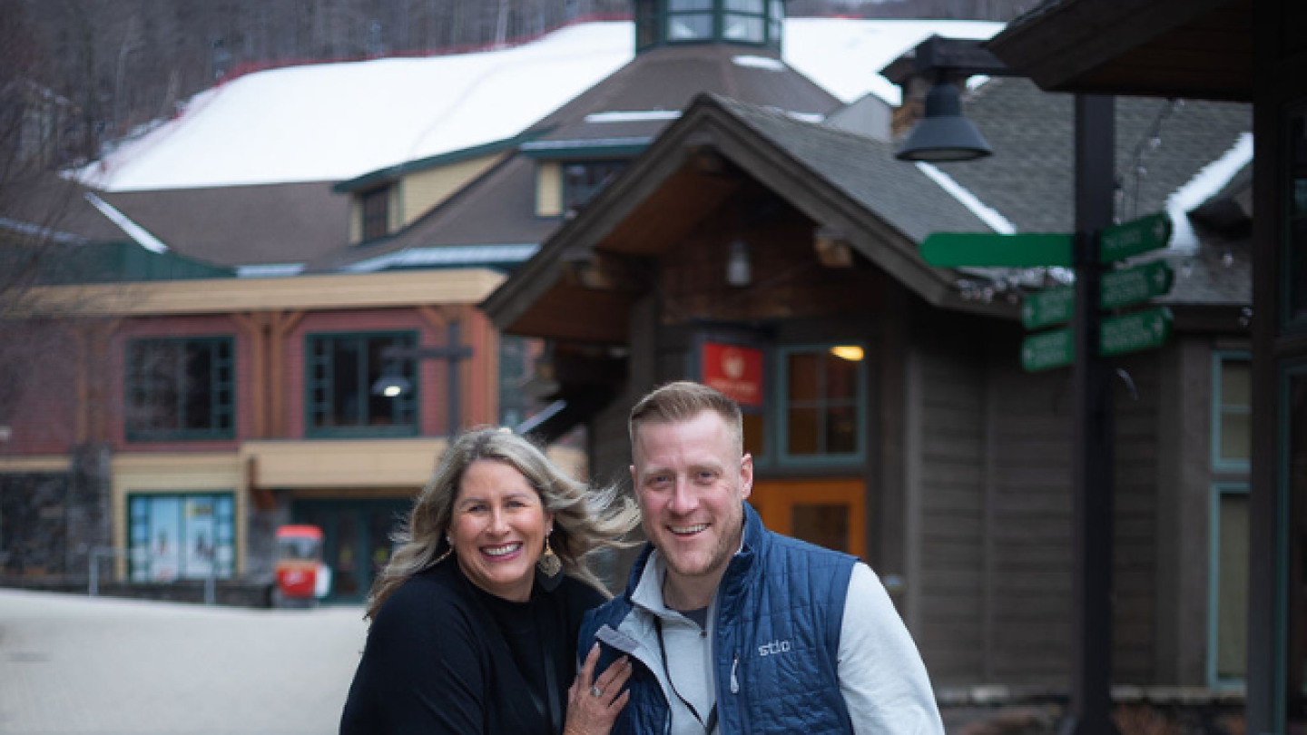 Two individuals pose and smile at the camera outside of the Lodge at Spruce Peak Resort