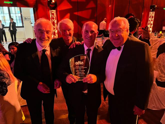 Four men wearing formal evening suits are pictured, with the third man from the left holding a crystal trophy